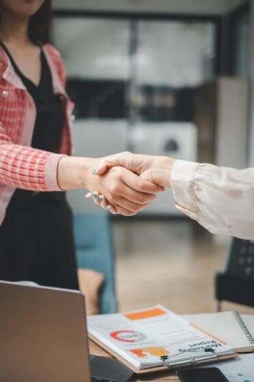 Two smiling businesswomen shaking hands in agreement during a successful office meeting, with a laptop on the table.